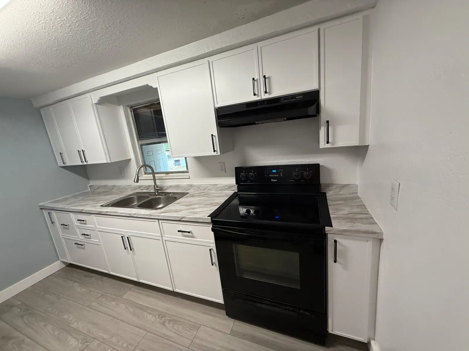 White and gray kitchen with upper and lower cabinets, sink, range, and marble look counters.